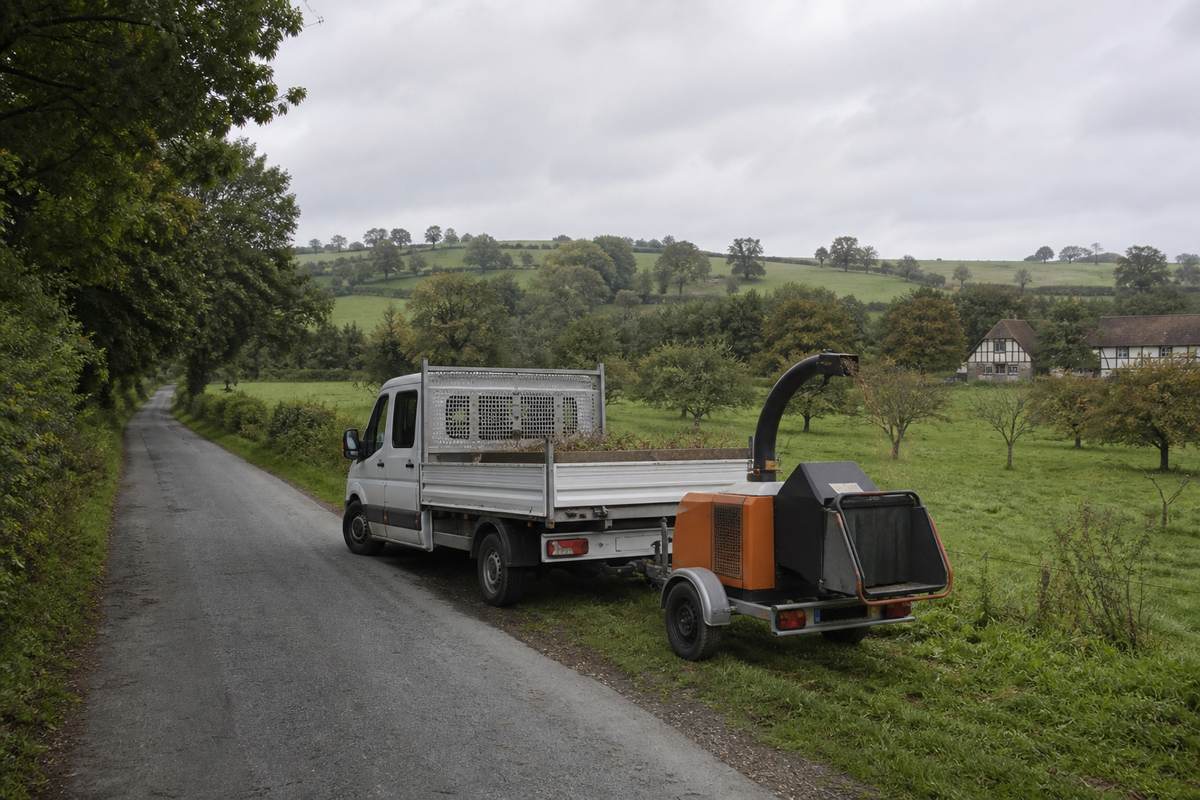 Camion et remorque de broyage stationnés au bord d'une route de campagne du Pays d'Auge, bordée de haies et de prairies à pommiers